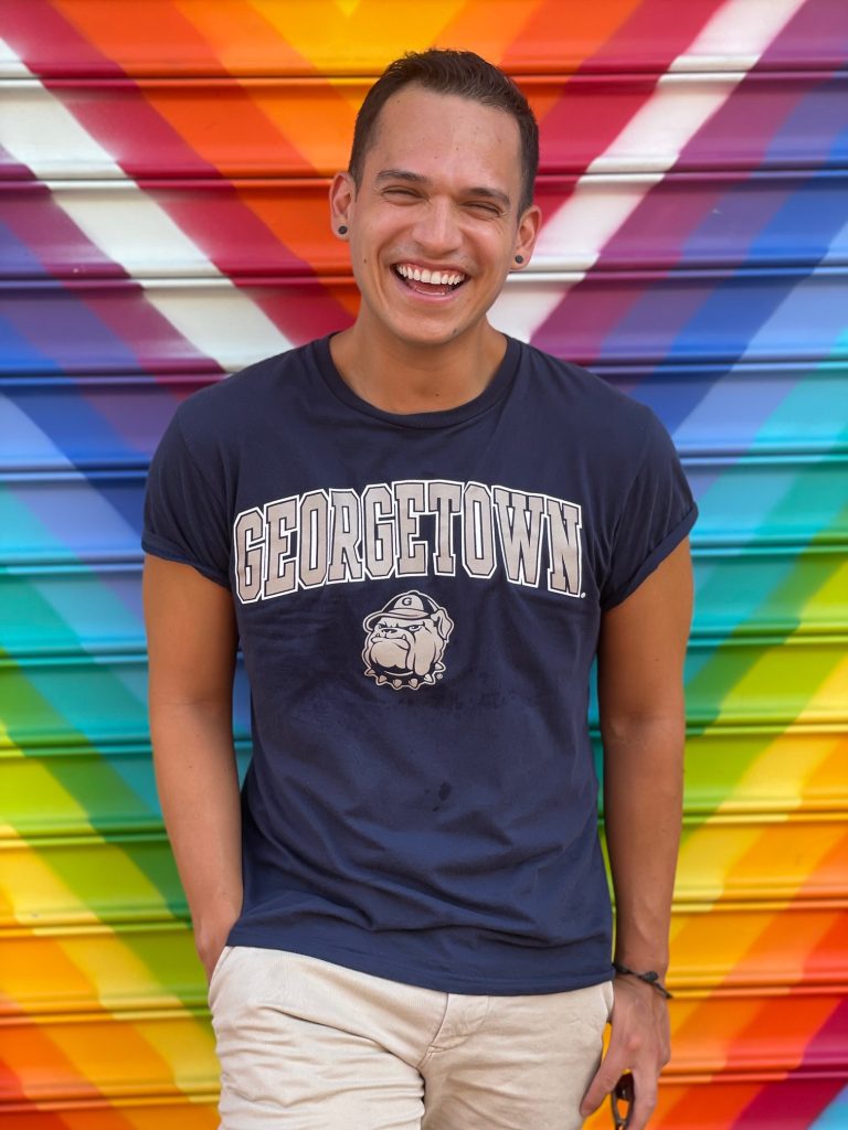 Photo of Brian in a Georgetown tee-shirt in front of a rainbow colored wall. 
